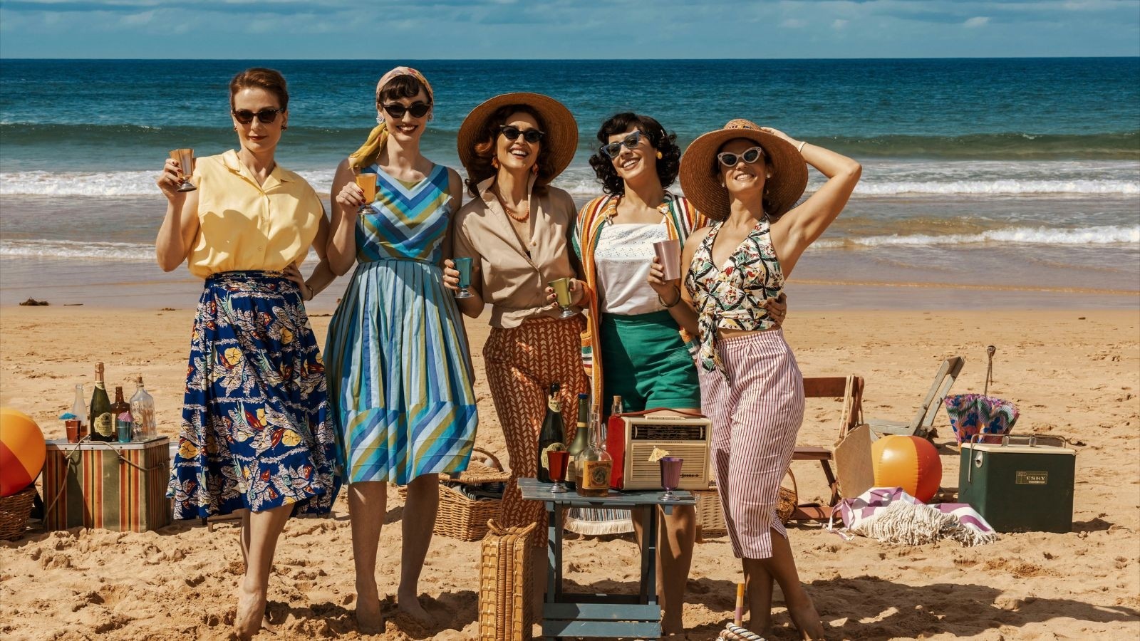 The cast of Ten Pound Poms stood on a beach in 50s clothing