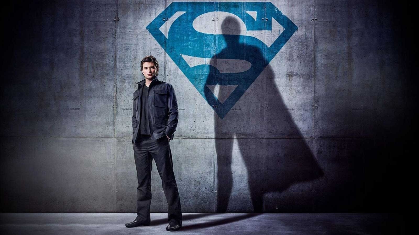 Tom Welling as Clark Kent in Smallville, stood in a dark room against a superman sign